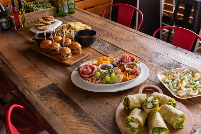 A wooden table set with sliders, a charcuterie platter, chicken wraps, devilled eggs and pasta salad